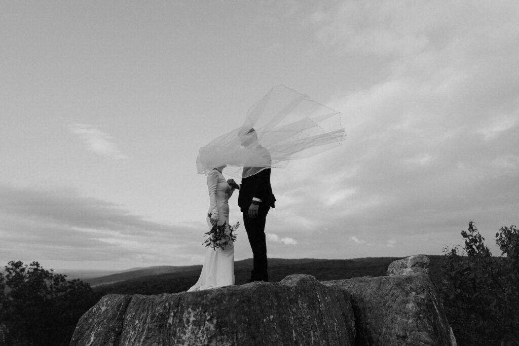 black and white photo of bride and groom on mountain with veil blowing in wind
