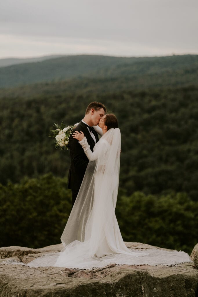 Bride and groom kiss on mountaintop with long cathedral veil