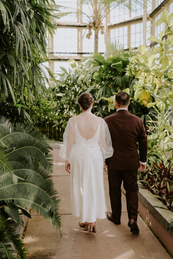 bride and groom walking around the rawlings conservatory during their baltimore elopement day surrounded by lush greenery and flowers