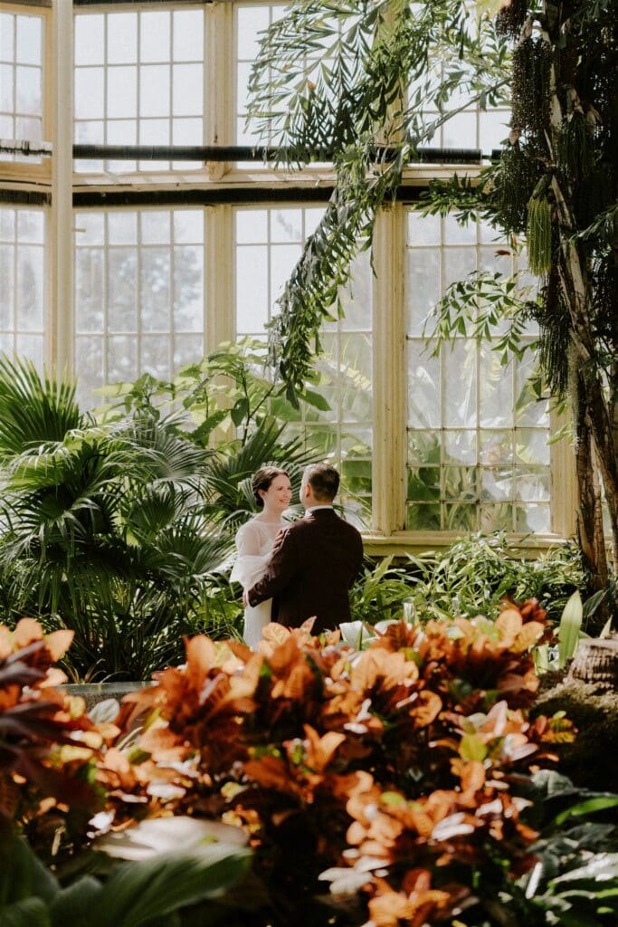 bride and groom walking around the rawlings conservatory during their baltimore elopement day surrounded by lush greenery and flowers